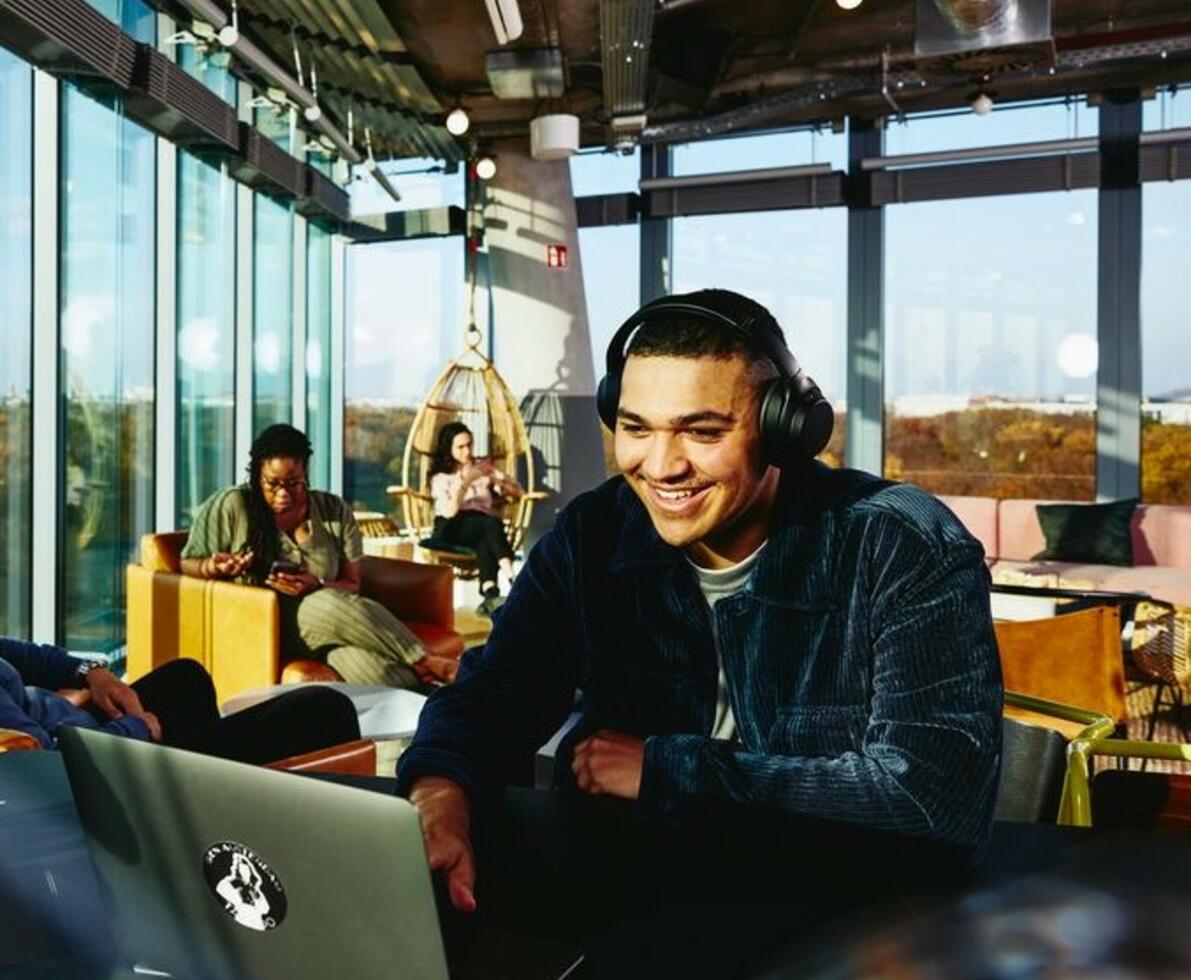 Smiling man with headphones and laptop sitting in a lounge environment.