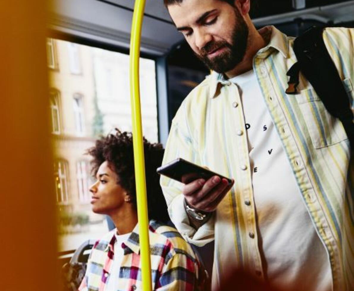Man on his phone standing in a bus. Woman sitting down in the background.
