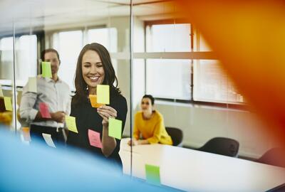 Two business women and a man in an office putting sticky notes on a window. Primary colors: yellow and blue.